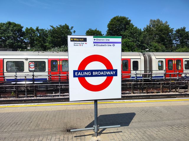 An Ealing Broadway roundel sign on a platform, with direction signs for the District and Elizabeth lines and a District line train behind