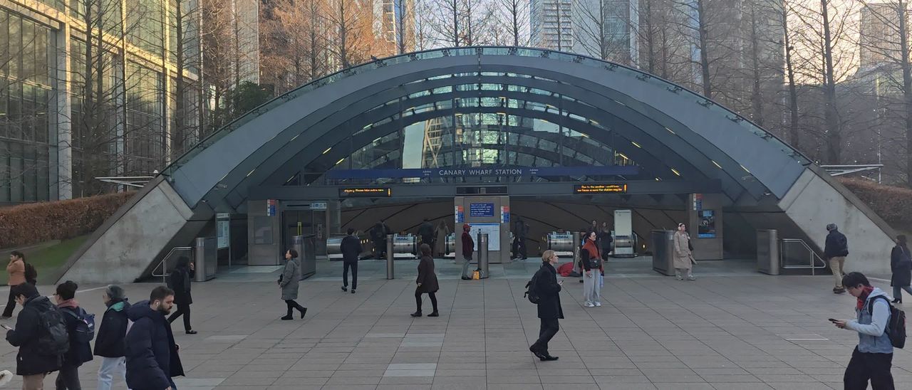 The curved glass canopy over the Jubilee line entrance at Canary Wharf, with people crossing the paved plaza in front