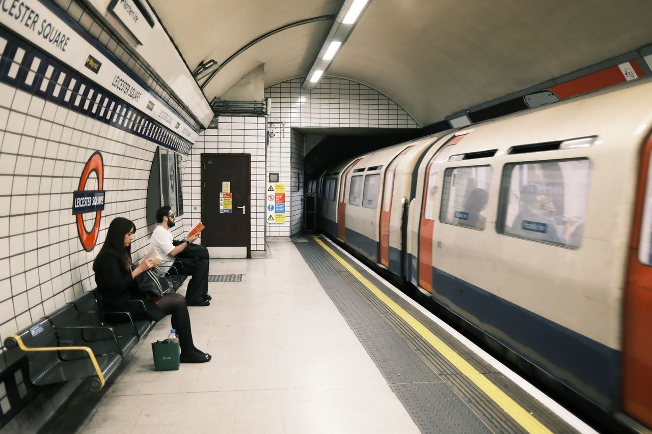 Leicester Square platform with a roundel on the tiled wall, people on a bench and a train in the station