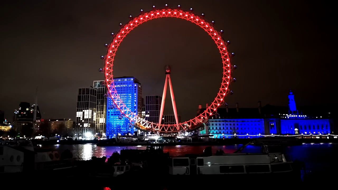 The London Eye lit red at night beside the river, with blue-lit buildings next to it