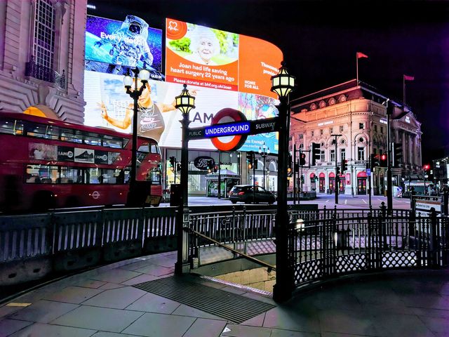 An Underground roundel on a lamp post above the railed stairway entrance at Piccadilly Circus at night, with lit advertising screens and a red bus behind