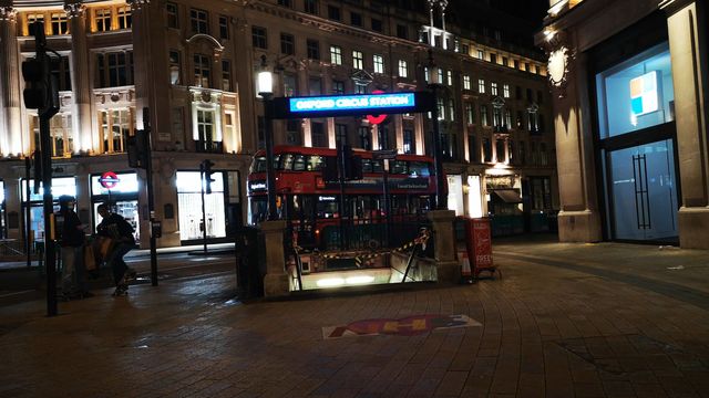 A lit Oxford Circus Station sign over railed stairs down to the Underground at night, with a red double-decker bus and lit buildings behind