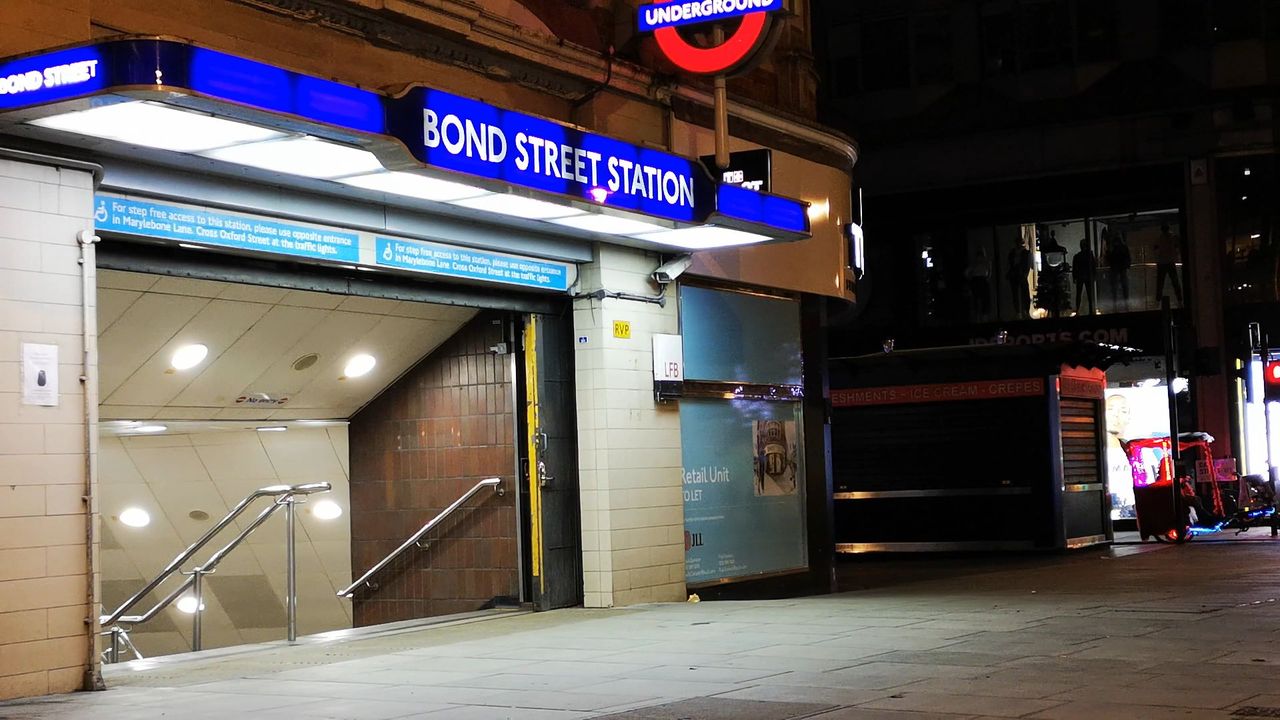 A lit Bond Street Station sign and Underground roundel over a stairway entrance on an empty street at night