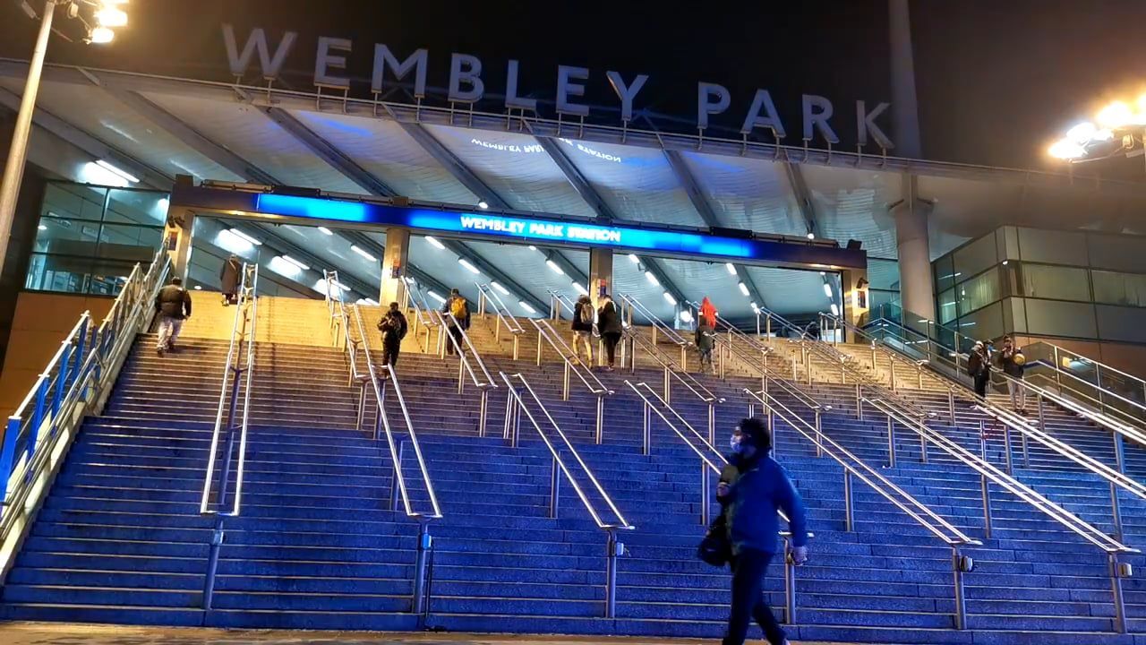 The blue-lit steps up to Wembley Park station at night, under a roof sign reading WEMBLEY PARK
