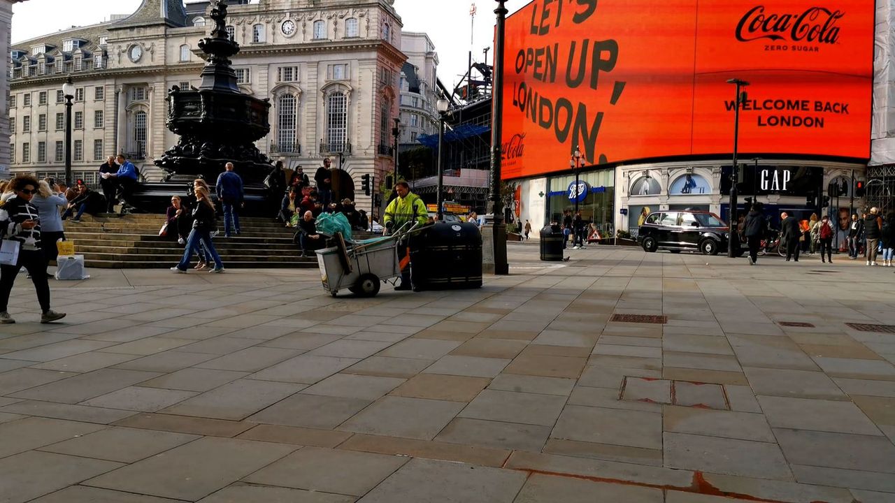 People crossing Piccadilly Circus by a statue on a stone base, with a large red digital screen on the buildings behind